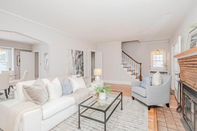 living area featuring arched walkways, light wood-style flooring, stairs, a brick fireplace, and plenty of natural light