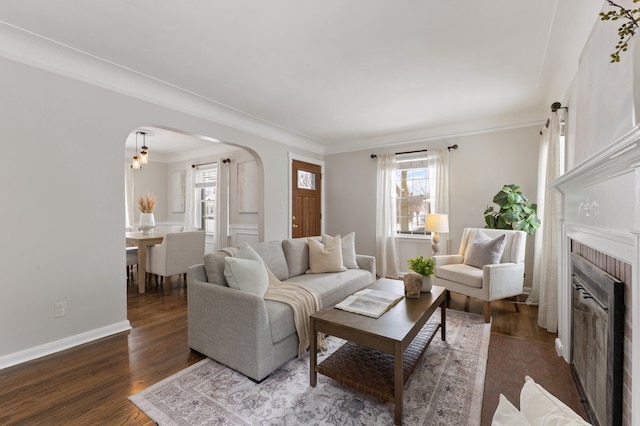 living area with arched walkways, dark wood finished floors, ornamental molding, a brick fireplace, and baseboards