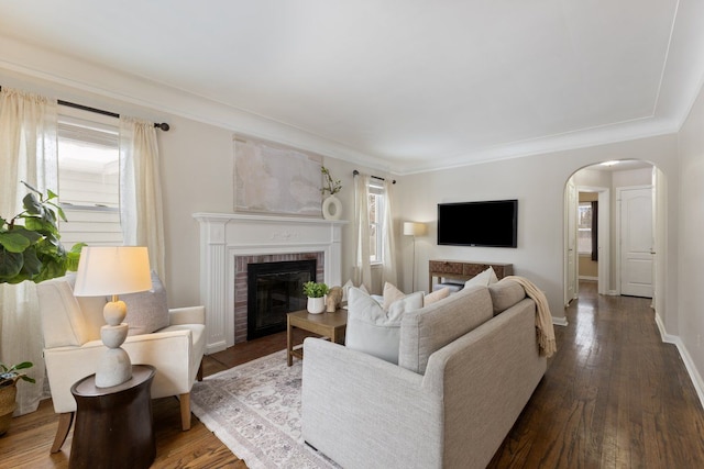 living area featuring baseboards, arched walkways, dark wood-style floors, crown molding, and a brick fireplace