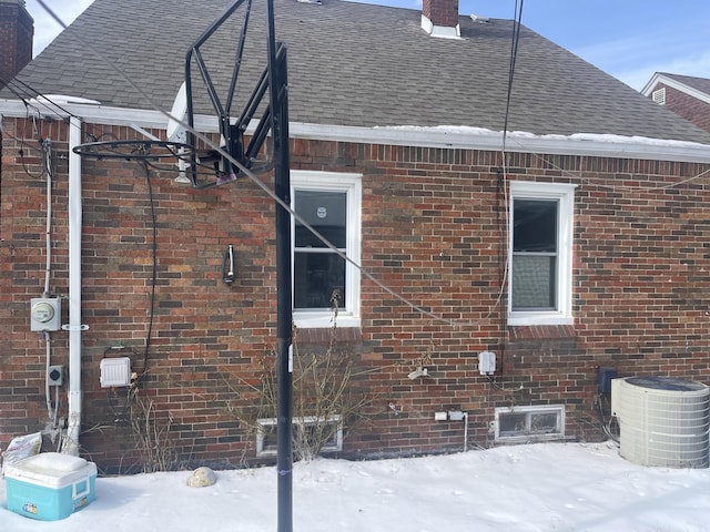 snow covered property featuring roof with shingles, brick siding, a chimney, and central air condition unit