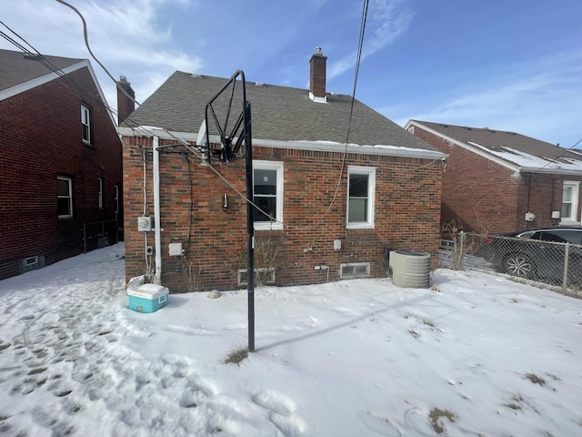 snow covered back of property featuring brick siding, a chimney, a shingled roof, and fence