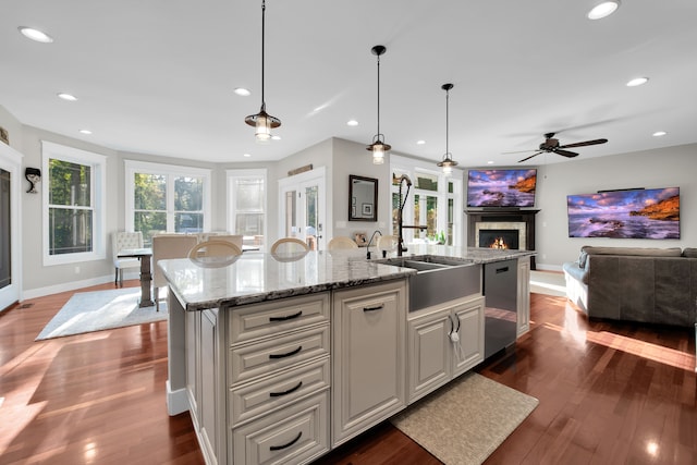 kitchen featuring a center island with sink, stainless steel dishwasher, open floor plan, light stone countertops, and a lit fireplace