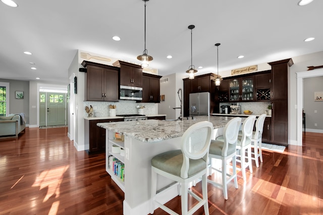 kitchen with decorative light fixtures, appliances with stainless steel finishes, glass insert cabinets, dark brown cabinetry, and a large island with sink