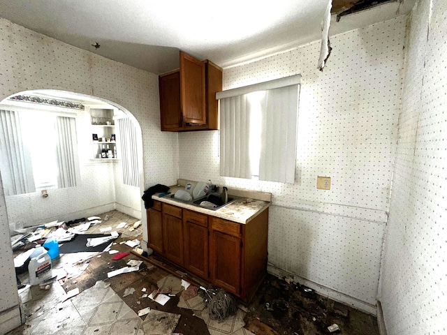 kitchen featuring arched walkways, light countertops, brown cabinets, and wallpapered walls