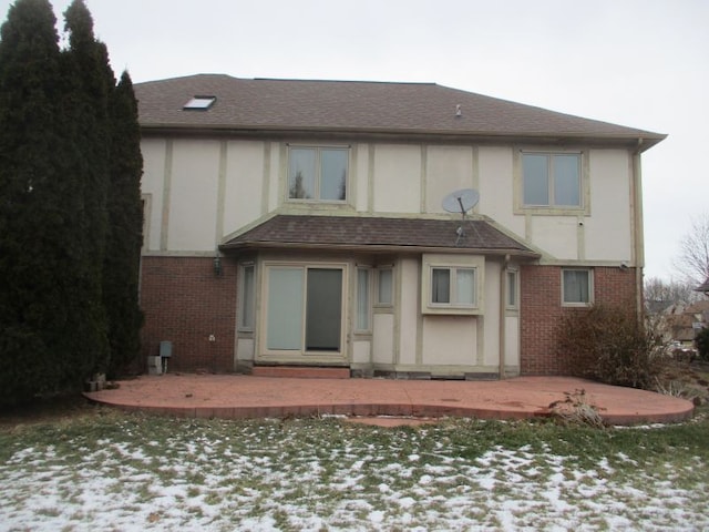 snow covered house featuring a patio area, brick siding, roof with shingles, and stucco siding