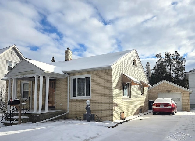 snow covered property featuring an outbuilding, brick siding, a chimney, and a detached garage