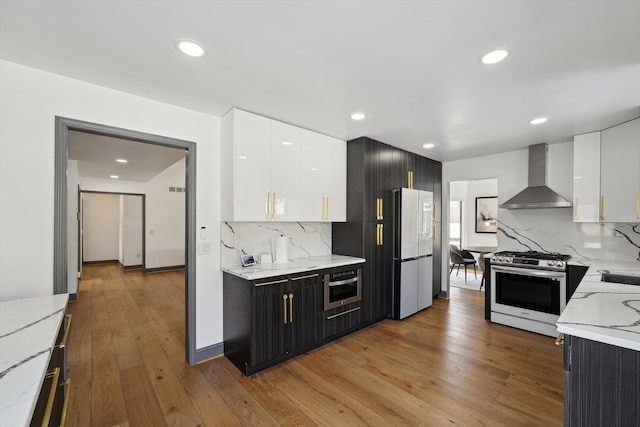 kitchen featuring white cabinets, wood-type flooring, light stone countertops, stainless steel appliances, and wall chimney range hood