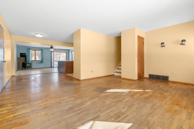 unfurnished living room with ceiling fan, light wood-style flooring, visible vents, stairway, and a wood stove