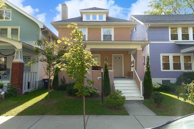 american foursquare style home featuring covered porch, a front yard, a chimney, and stucco siding