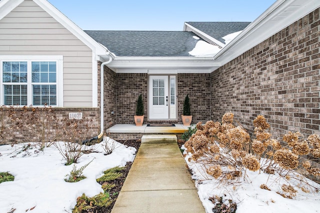 snow covered property entrance with a shingled roof and brick siding