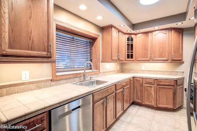 kitchen with light tile patterned floors, tile counters, stainless steel dishwasher, glass insert cabinets, and a sink