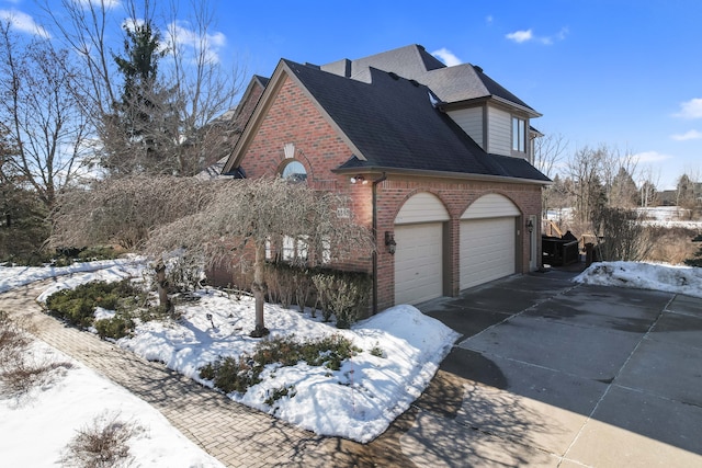 view of snowy exterior with a shingled roof, brick siding, driveway, and a garage