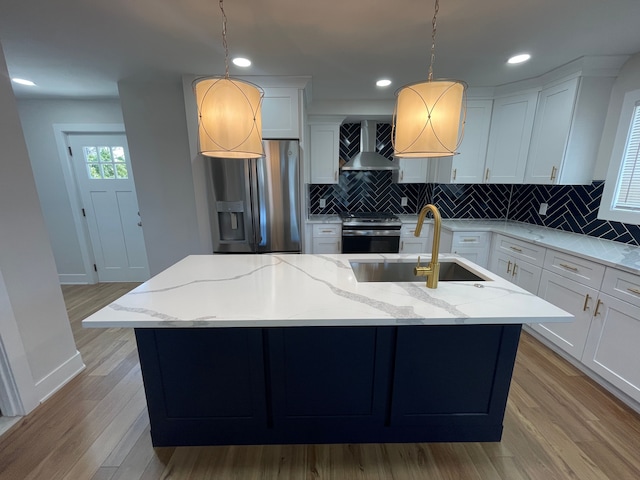 kitchen with a kitchen island with sink, stainless steel appliances, a sink, hanging light fixtures, and wall chimney range hood