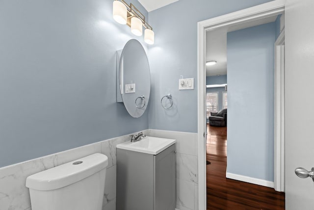 half bathroom featuring toilet, a wainscoted wall, wood finished floors, vanity, and tile walls