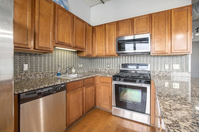 kitchen featuring light wood finished floors, appliances with stainless steel finishes, brown cabinetry, a sink, and dark stone counters