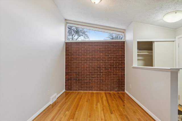 empty room with light wood-type flooring, visible vents, a textured ceiling, brick wall, and baseboards