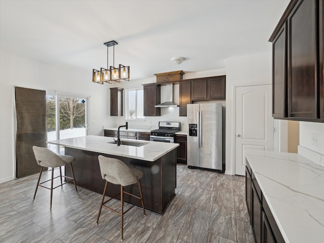 kitchen featuring appliances with stainless steel finishes, a sink, dark brown cabinets, light stone countertops, and wall chimney exhaust hood