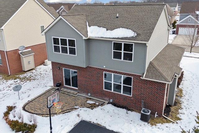 snow covered house featuring cooling unit, brick siding, a patio, and roof with shingles