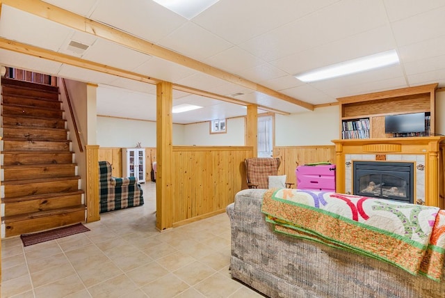 living room featuring wooden walls, a drop ceiling, a glass covered fireplace, wainscoting, and stairway