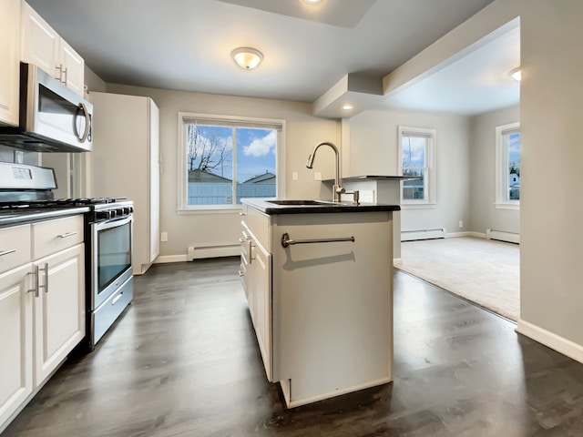 kitchen featuring an island with sink, dark countertops, baseboard heating, stainless steel appliances, and white cabinetry