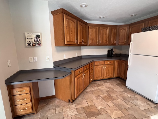 kitchen featuring dark countertops, brown cabinets, built in desk, and freestanding refrigerator