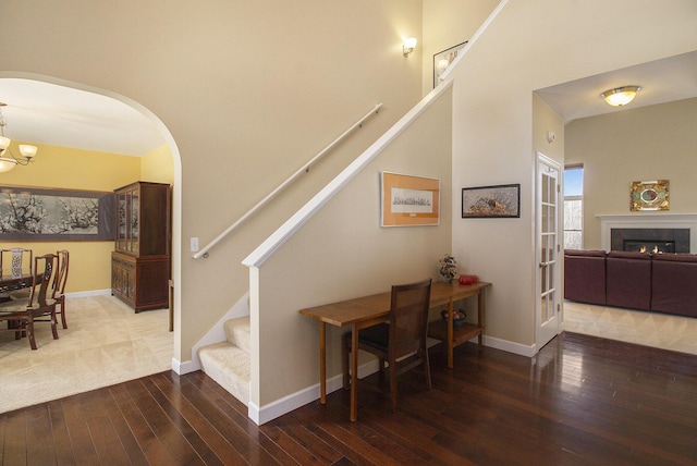 stairway featuring a warm lit fireplace, arched walkways, baseboards, wood-type flooring, and a notable chandelier