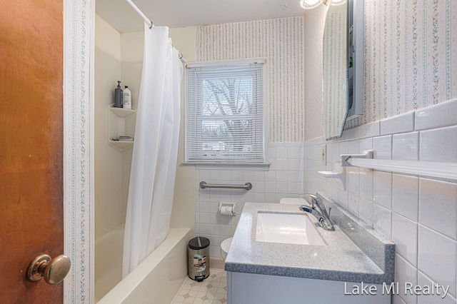 bathroom with wallpapered walls, wainscoting, shower / bathtub combination with curtain, vanity, and tile walls