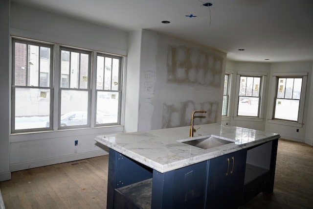 kitchen with light stone counters, a kitchen island with sink, a sink, blue cabinetry, and plenty of natural light