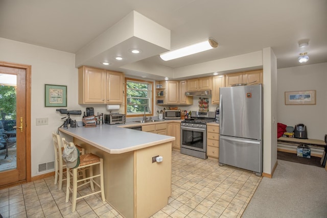 kitchen featuring a toaster, a peninsula, stainless steel appliances, light brown cabinetry, and under cabinet range hood