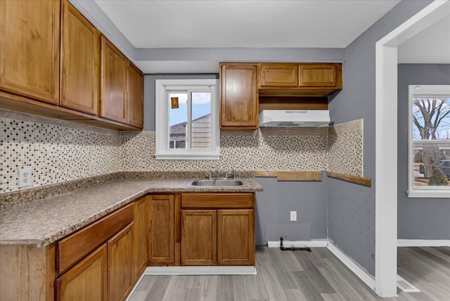 kitchen featuring under cabinet range hood, a sink, baseboards, brown cabinets, and tasteful backsplash