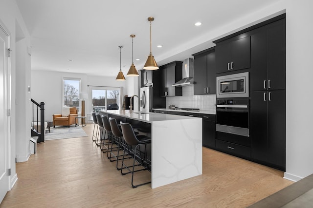 kitchen featuring stainless steel appliances, light countertops, a kitchen island with sink, wall chimney range hood, and dark cabinetry