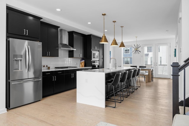 kitchen with an island with sink, appliances with stainless steel finishes, hanging light fixtures, dark cabinetry, and wall chimney range hood