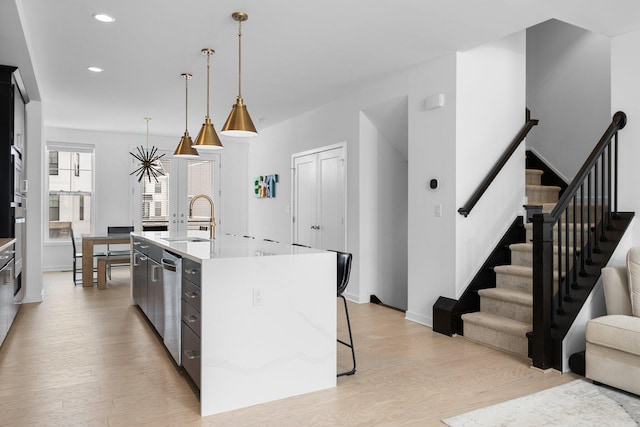 kitchen featuring a center island with sink, hanging light fixtures, a sink, light wood-type flooring, and dishwasher