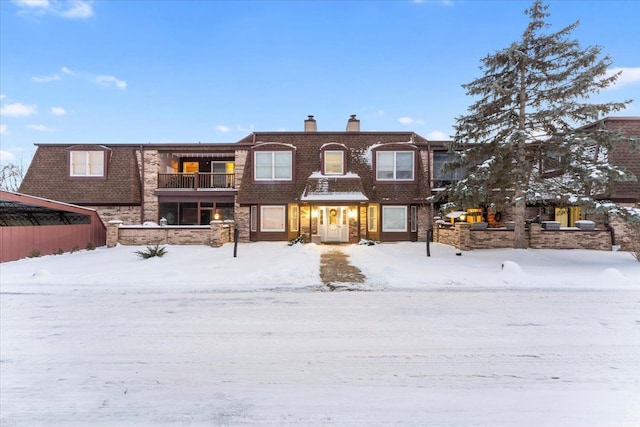 snow covered rear of property with a balcony, a chimney, mansard roof, and brick siding