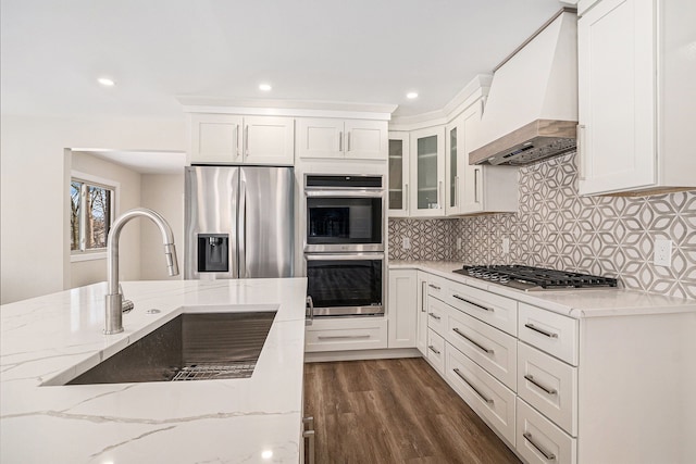 kitchen featuring glass insert cabinets, appliances with stainless steel finishes, light stone counters, custom exhaust hood, and a sink