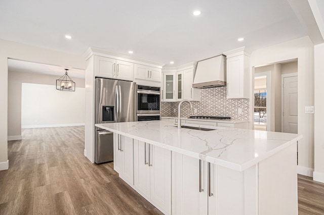 kitchen with premium range hood, a kitchen island with sink, glass insert cabinets, and appliances with stainless steel finishes