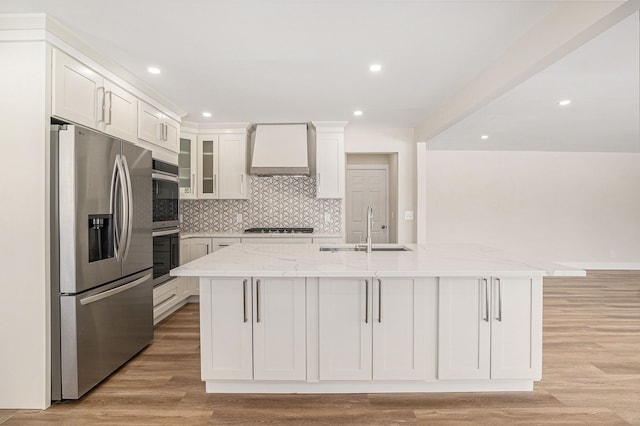 kitchen featuring stainless steel appliances, an island with sink, glass insert cabinets, and custom range hood