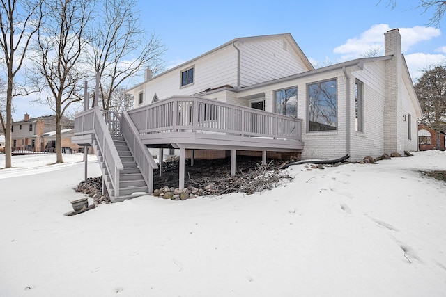 snow covered back of property featuring stairs, a chimney, a deck, and brick siding