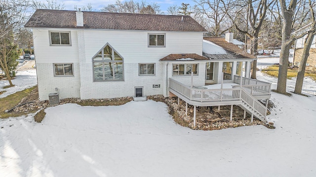 snow covered property with a deck, cooling unit, brick siding, and a chimney