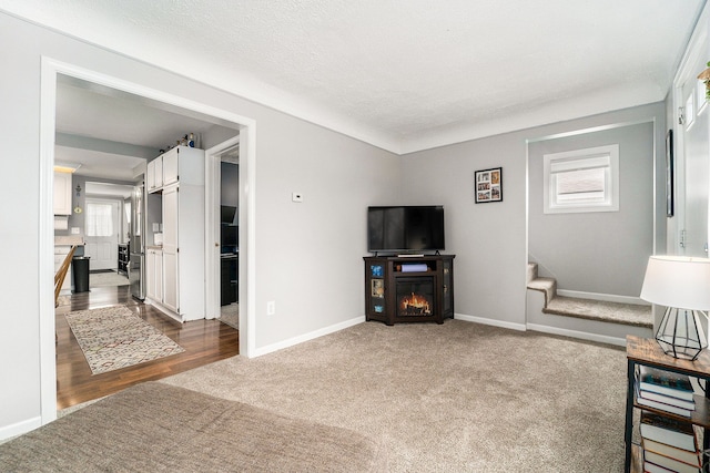 living room with a warm lit fireplace, a textured ceiling, baseboards, stairs, and dark carpet