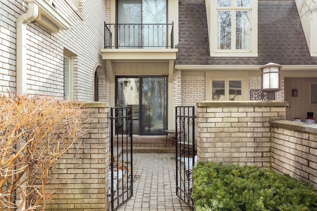 property entrance featuring mansard roof, a balcony, brick siding, roof with shingles, and a gate