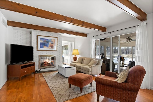 living room featuring a brick fireplace, beam ceiling, visible vents, and wood finished floors