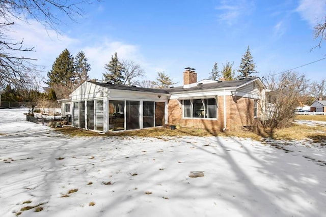 snow covered rear of property with brick siding, a chimney, and a sunroom