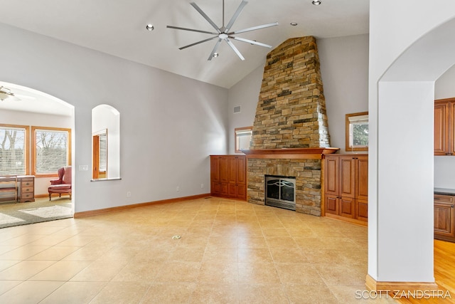 unfurnished living room with arched walkways, high vaulted ceiling, a stone fireplace, a ceiling fan, and baseboards