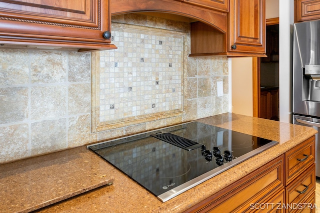 kitchen with stainless steel fridge, decorative backsplash, light stone counters, brown cabinets, and black electric cooktop