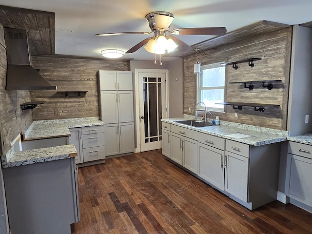 kitchen featuring light stone counters, a sink, a ceiling fan, wall chimney exhaust hood, and dark wood finished floors