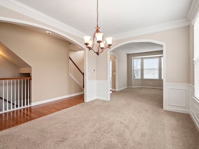 carpeted spare room featuring arched walkways, a notable chandelier, wainscoting, and crown molding