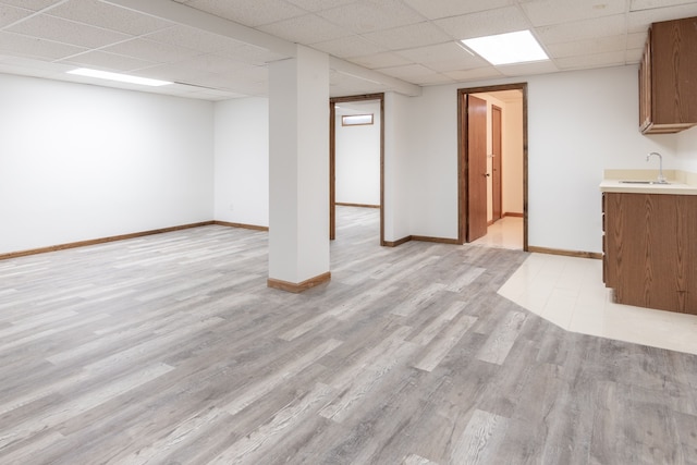 finished below grade area featuring light wood-style flooring, baseboards, a drop ceiling, and a sink