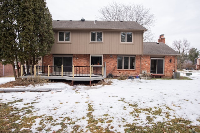 snow covered rear of property with a deck, brick siding, a chimney, and central air condition unit