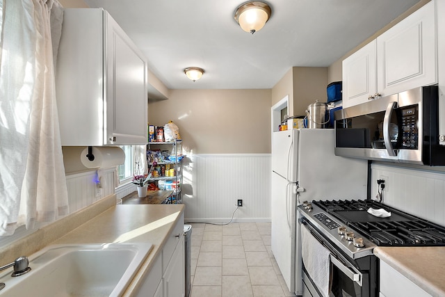 kitchen with light tile patterned floors, a wainscoted wall, stainless steel appliances, light countertops, and white cabinetry